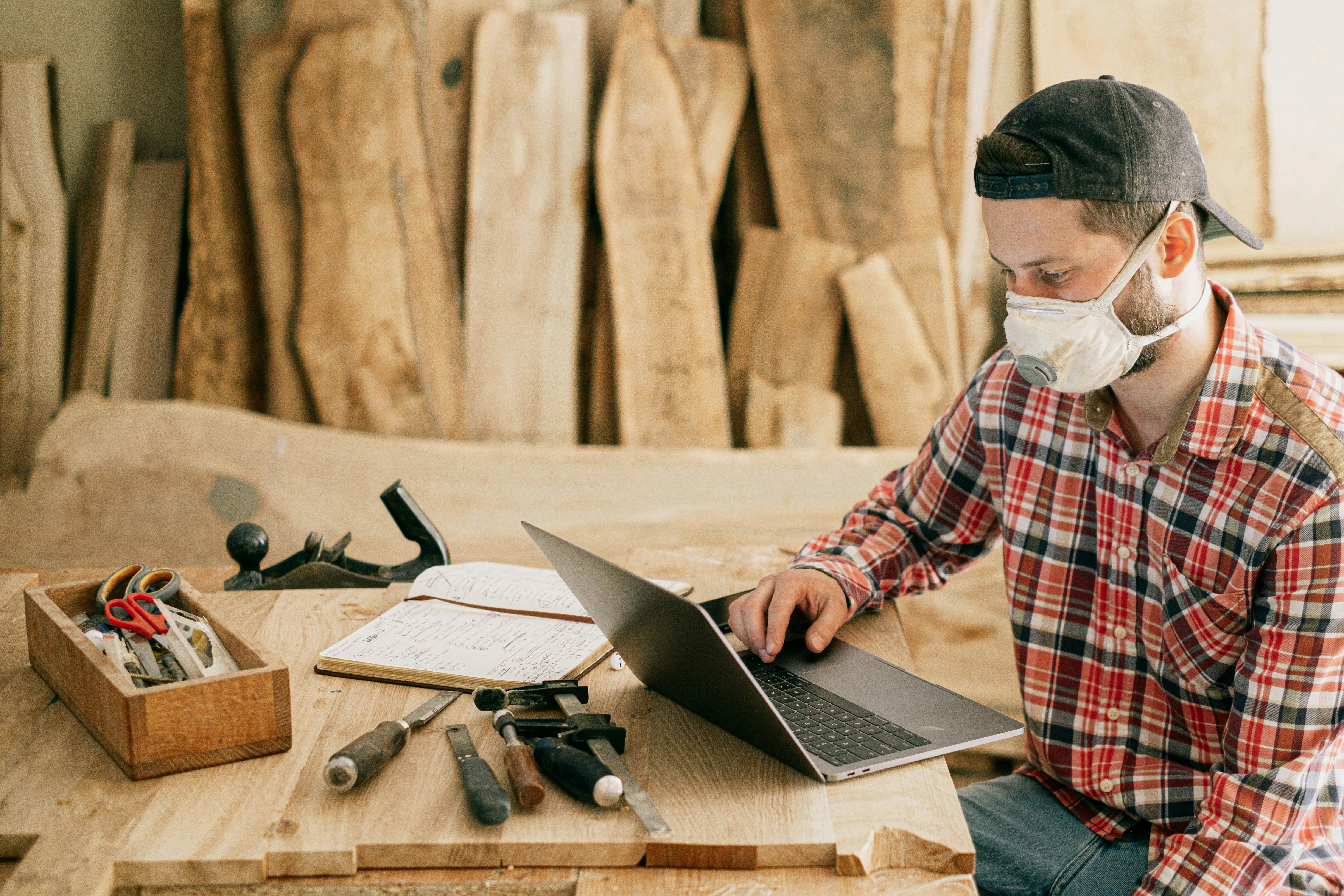 Man in a dust mask sits at a carpenter's work table and looks at laptop 